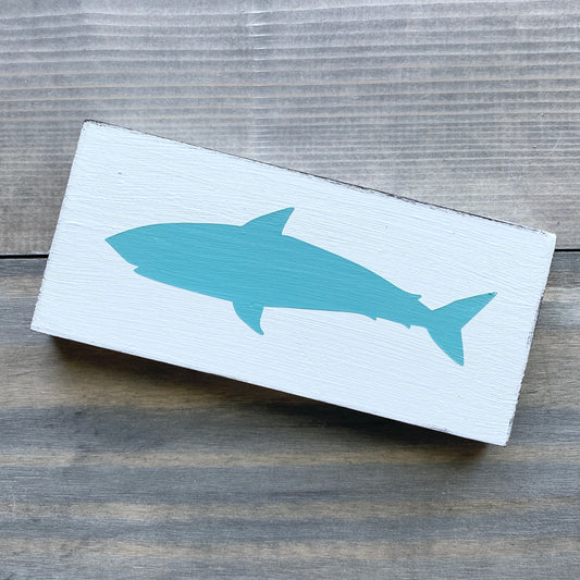 A rectangular white wooden sign featuring a painted aqua shark silhouette, displayed on a rustic gray wood plank surface.