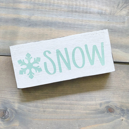 A rectangular white wooden sign with the word 'SNOW' in mint and a snowflake icon, photographed on a rustic gray-brown wood plank surface.