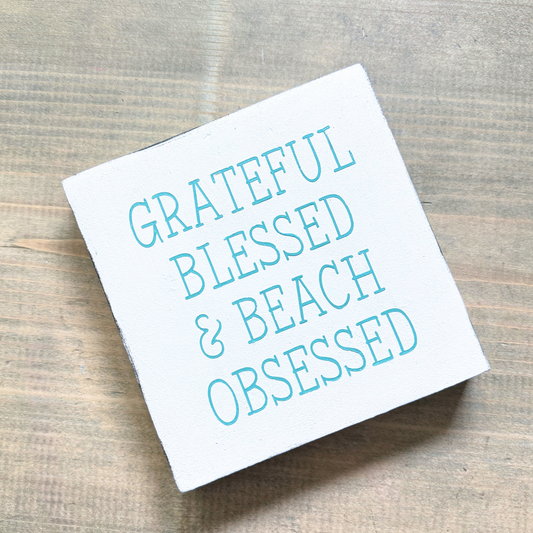 A square white decorative sign with the phrase “GRATEFUL BLESSED & BEACH OBSESSED” in aqua lettering, photographed on a rustic wood surface.