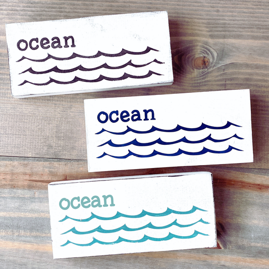 Three rectangular white wooden coasters with the word 'ocean' and wave illustrations, shown on a rustic wood surface. The coasters are white with dark purple, navy blue, and teal lettering and wave patterns.