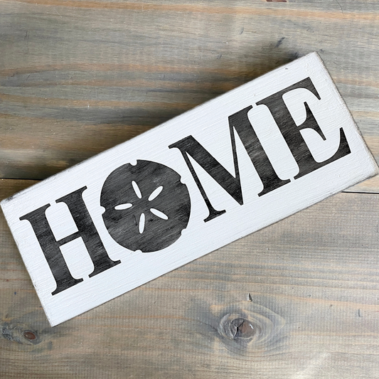 A rectangular white wooden sign with the word "HOME" in large black letters, where the letter O is replaced by a black sand dollar graphic. The sign is photographed on a rustic gray-brown wooden surface.