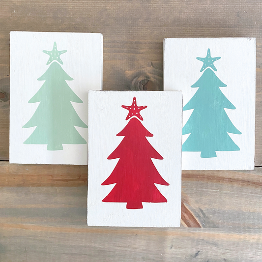 Three hand-painted wooden Christmas signs with tree silhouettes and starfish toppers, displayed on a rustic wooden surface. The center sign is white with a red tree and star, while the signs behind it are also white with mint and aqua trees.