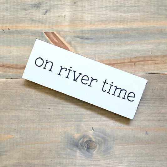 A white rectangular sign with the phrase 'on river time' in black lettering, photographed on a rustic light brown wood plank surface.