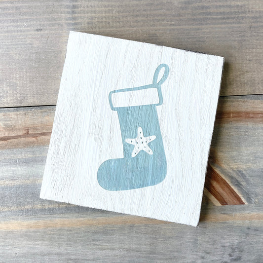A square white wooden sign with a painted sage green Christmas stocking and a white starfish, photographed on a rustic wood plank surface.