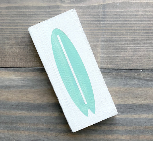 A rectangular white wooden sign featuring a painted mint green surfboard design, placed diagonally on a dark wood plank surface.