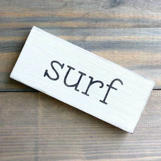 A rectangular white wooden sign with the word 'surf' in black lowercase placed diagonally on a rustic brown wood plank surface.