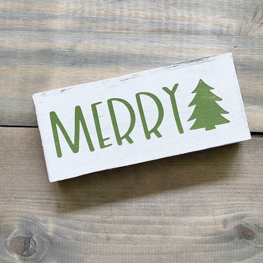 A rectangular white wooden sign with 'MERRY' in green lettering and a green Christmas tree icon, photographed on a rustic wood plank surface.