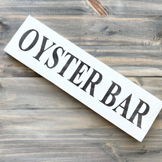 A rustic white wooden sign with black text reading 'OYSTER BAR', placed diagonally on a weathered gray-brown wooden plank surface.