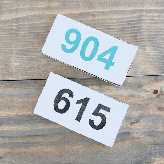 Two rectangular white house number signs with rustic edges, one displaying '904' in teal and the other '615' in black, arranged diagonally on a weathered wood plank surface.