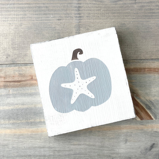 A square white wooden sign featuring a painted gray pumpkin with a white starfish on the front, photographed on a rustic gray wood plank surface.