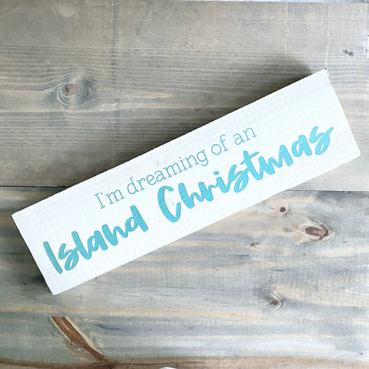 A white rectangular wooden sign with the phrase “I’m dreaming of an Island Christmas” in aqua blue lettering, placed diagonally on a rustic gray wood plank surface.