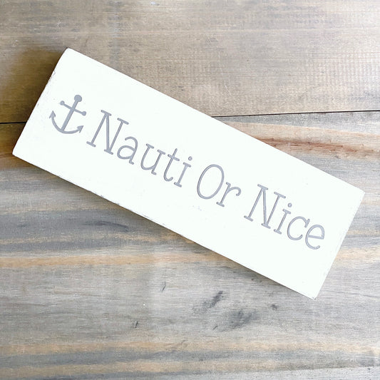 A rectangular white wooden sign with a gray anchor and the words 'Nauti Or Nice' in gray script, placed diagonally on a rustic gray-brown wooden surface.