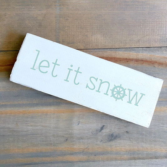 A rectangular white wooden sign with the phrase 'let it snow' in a light mint green font, featuring a snowflake icon where the letter 'o' appears. The sign is photographed at an angle on a rustic, weathered brown wooden surface.