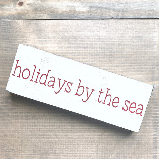 white sign with red lettering saying holidays by the sea placed on a wooden surface
