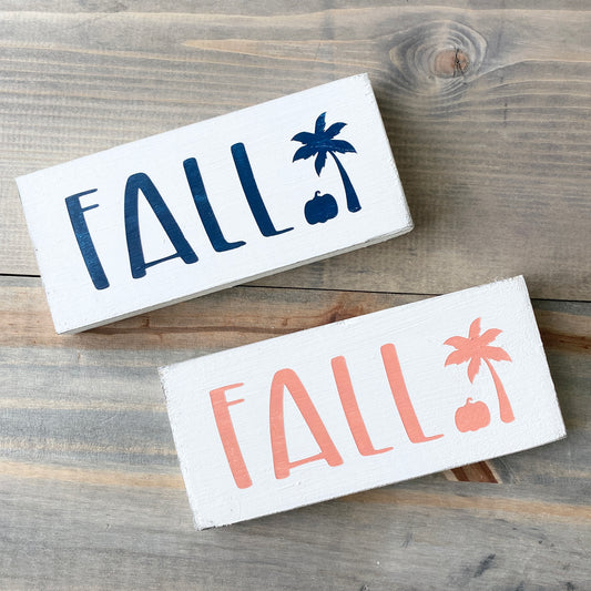 Two wooden signs with 'FALL' text and palm tree and pumpkin design on a wooden surface