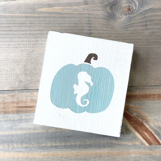 A square white wooden sign featuring a painted sage green pumpkin with a white seahorse silhouette in the center and a dark brown stem, displayed on a rustic wood plank surface.