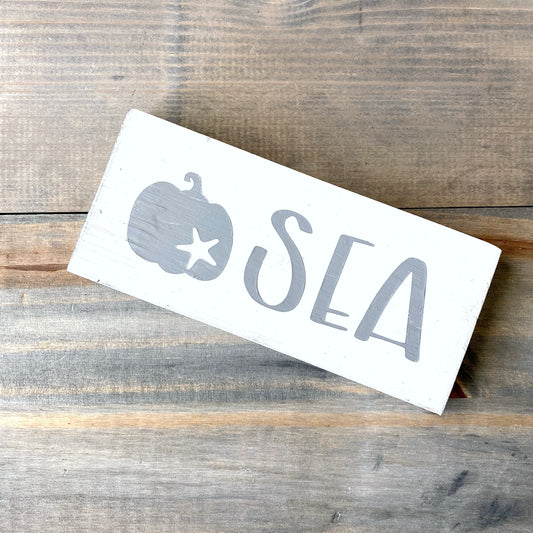 A rectangular white wooden sign with a gray painted pumpkin with starfish and the word 'SEA' in large gray letters, placed diagonally on a rustic wood plank surface.