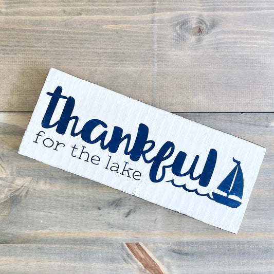 Thankful for the lake sign in white with navy lettering and sailboat and wave graphic. Laying on a rustic wood background