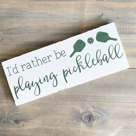 A rectangular white wooden sign with rustic edges, printed with the phrase 'I'd rather be playing pickleball' in dark green lettering, alongside simple green pickleball paddle and ball illustrations. The sign is photographed at an angle on a weathered gray-brown wooden surface.