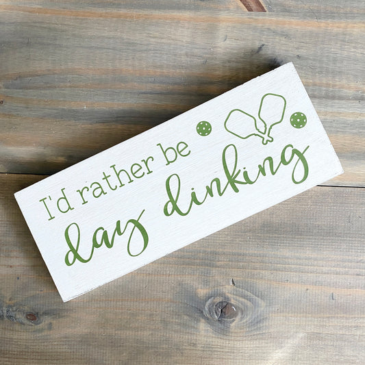 A wooden sign with a white background, featuring the phrase 'I'd Rather Be Day Dinking' in green text, along with small green illustrations of pickle balls and paddles