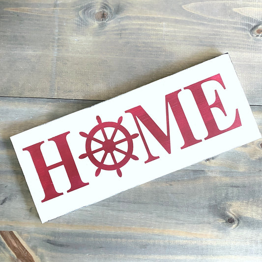 A rectangular white wooden sign with the word "HOME" in red, where the letter O is replaced by a red ship's wheel, photographed on a rustic gray-brown wood plank surface.