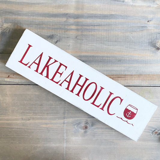 A white rectangular wooden sign with the word 'LAKEAHOLIC' printed in dark red serif letters and a small anchor-in-a-glass icon at the end, photographed on a rustic gray-brown wood plank surface.