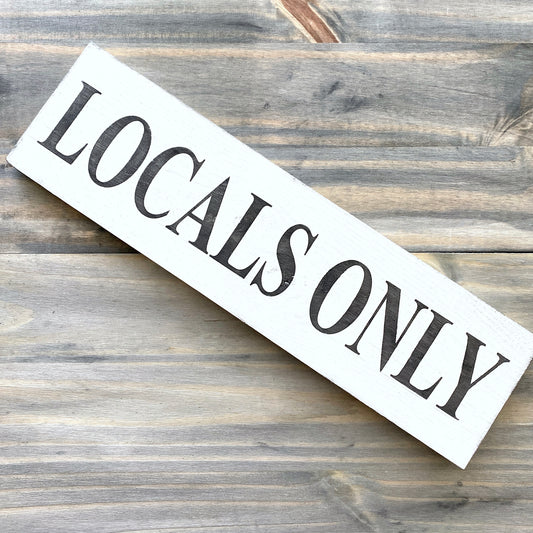 A rectangular white wooden sign with the words 'LOCALS ONLY' in large black serif lettering, placed diagonally on a rustic gray-brown wood plank surface.