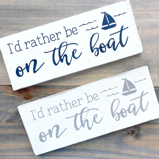 Two wooden signs with 'I'd rather be on the boat' text and sailboat graphics on a wooden surface.