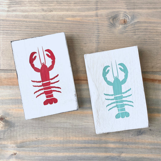 Two small rectangular white wooden signs with painted lobster illustrations, one in red and one in aqua displayed on a rustic wood plank surface.