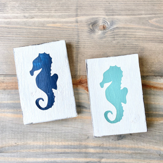 Seahorse Sign Coastal home decor and inspiration for the beach lover, gifts from Anchoredsouldesigns Two wooden signs with a seahorse design, one painted navy and the other in aqua, placed on a wooden surface.