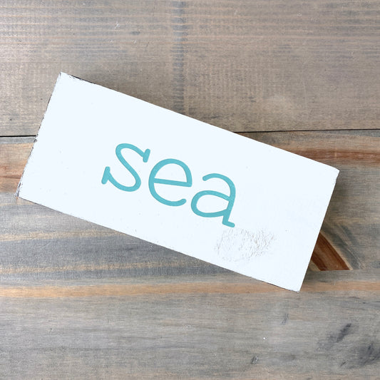 A white rectangular wooden sign with the word 'sea' painted in aqua lowercase letters, photographed on a rustic gray-brown wooden surface.