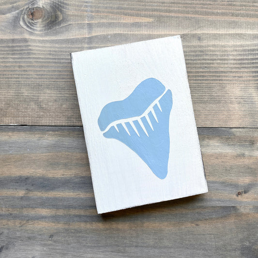 A rectangular white wooden block or sign featuring a large light blue shark tooth graphic, photographed from above on a rustic dark wood plank surface.