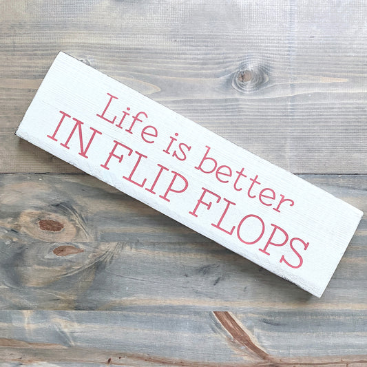 A rustic white wooden sign with pink text reading 'Life is better IN FLIP FLOPS' on a weathered gray wood plank background.