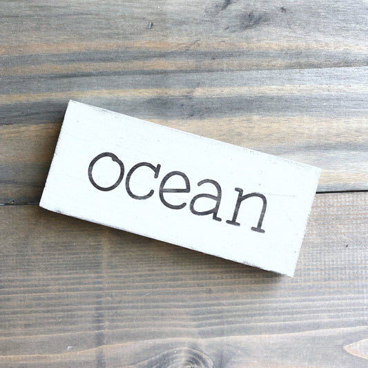 A rectangular white wooden sign with the word 'ocean' in black lowercase script, photographed on a rustic dark wood plank surface.