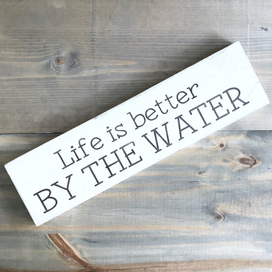A rectangular white wooden sign with the phrase 'Life is better BY THE WATER' in dark gray lettering, photographed diagonally on a rustic gray-brown wood plank surface.