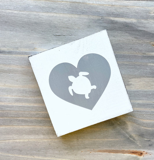 A square white wooden sign with a gray heart design containing a white sea turtle silhouette, placed on a rustic gray wood surface.