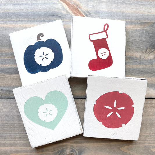Handcrafted sand dollar wood signs in four seasonal colorways including a navy pumpkin, red Christmas stocking, mint heart, and pink sand dollar representing fall, Christmas, Valentine's Day, and everyday coastal decor