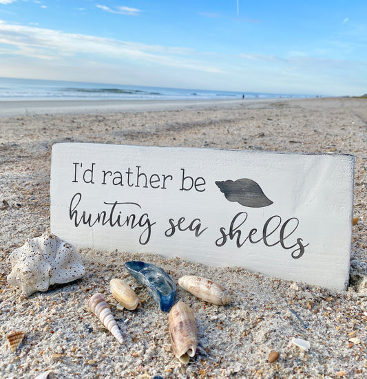 I'd Rather be hunting seashells Sign amidst a collection of shells laying on the beach: Memories of Beach Adventures