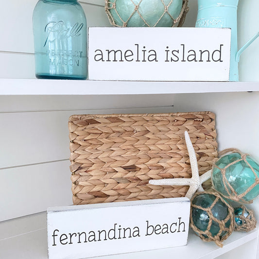 Handmade small location word signs reading Amelia Island and Fernandina Beach displayed on a coastal shelf with sea glass floats, starfish, and mason jars, personalized housewarming gifts for beach house owners
