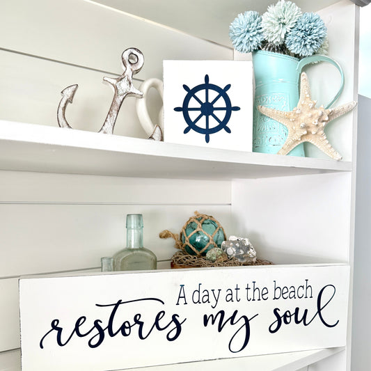 Handmade wood sign reading A Day at the Beach Restores My Soul styled on a shelf with a ship wheel and coastal decor