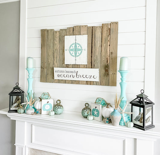 Coastal fall mantle with seafoam and white pumpkins, aqua candlesticks, black lanterns, glass floats, sand dollar and starfish signs, and an Autumn Leaves and Ocean Breeze sign from Anchored Soul Designs on a white shiplap wall