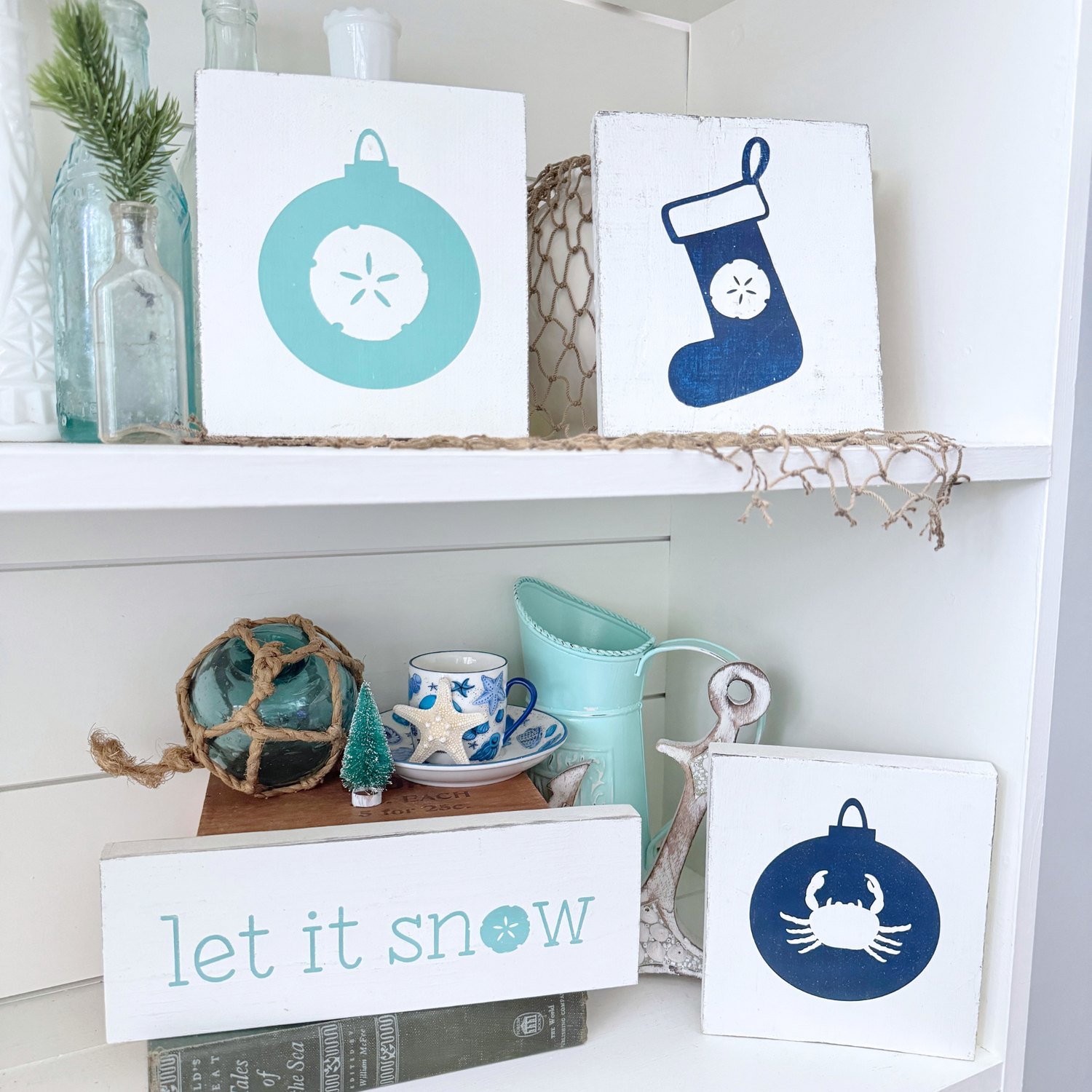 Handcrafted coastal Christmas signs including a sand dollar ornament, sand dollar stocking, crab ornament, and a "let it snow" sign styled on a white shiplap shelf with sea glass bottles and starfish accents