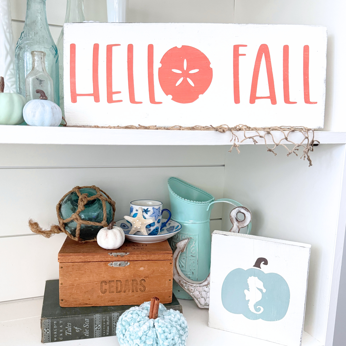 Coastal fall decor vignette featuring a "Hello Fall" sand dollar wood sign, seahorse pumpkin sign, white pumpkins, sea glass bottles, and a fishing float styled on a white shiplap shelf