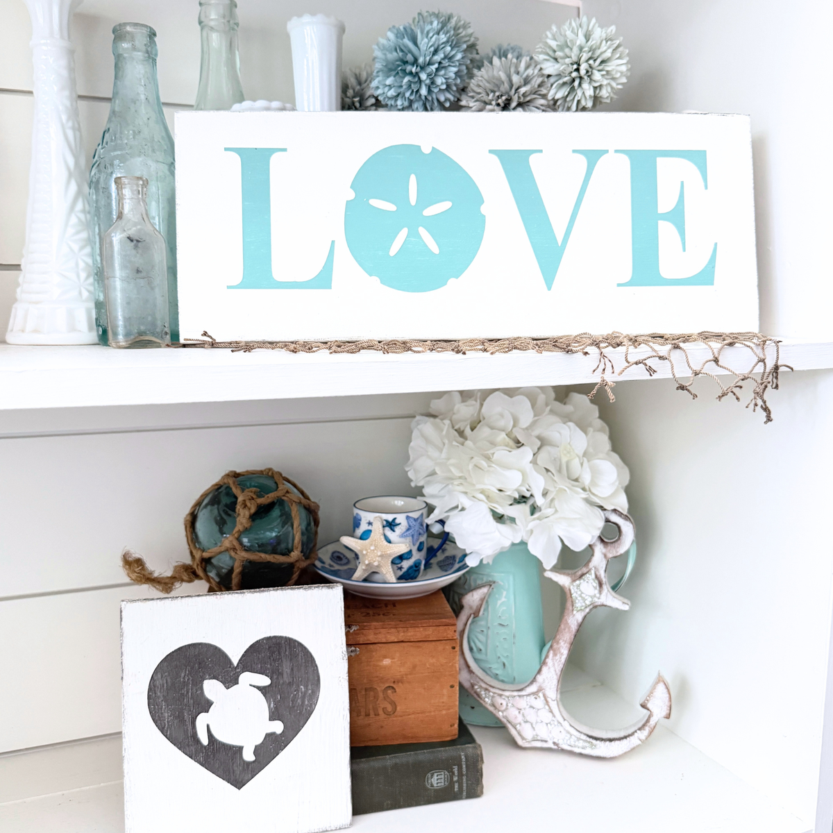 Coastal Valentine's Day decor featuring a sand dollar LOVE wood sign and sea turtle heart sign styled on a white shiplap shelf with sea glass bottles, hydrangeas, and an anchor accent
