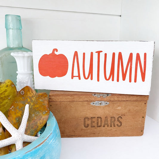 Wooden box with autumn-themed sign with pumpkin and 'AUTUMN' text, and decorative items on a white surface.