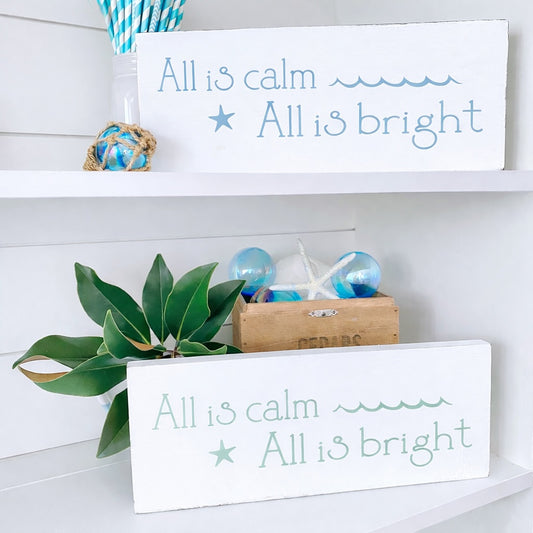 Decorative signs with 'All is calm, All is bright' text on a shelf with plants and decor items.