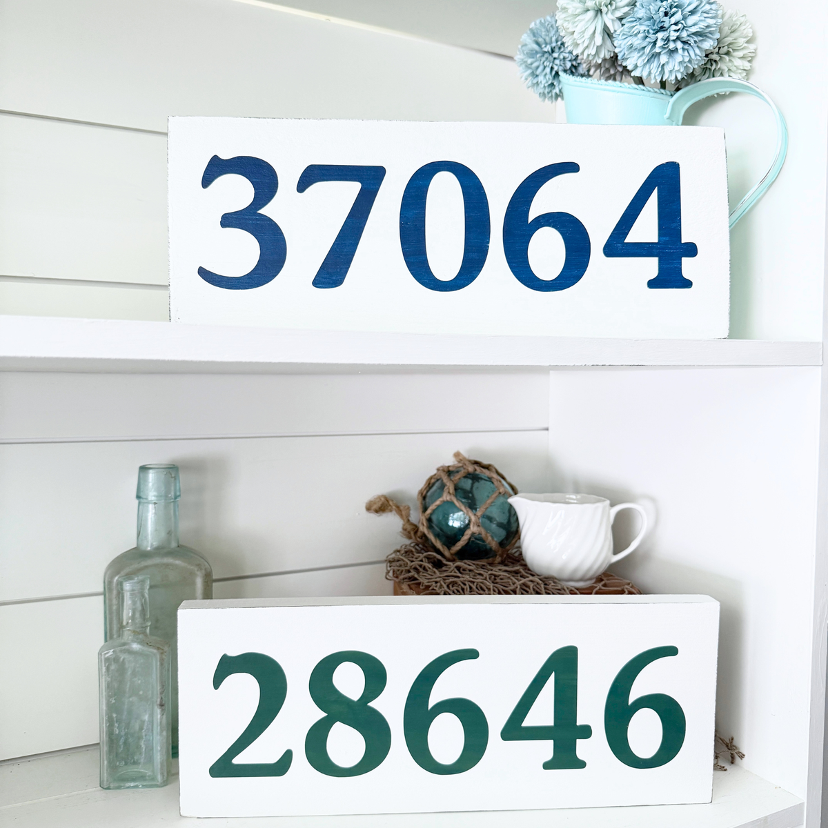 Two white wooden house numbers signs displayed on white shelves. The top sign reads '37064' in navy blue, and the bottom sign reads '28646' in dark green.