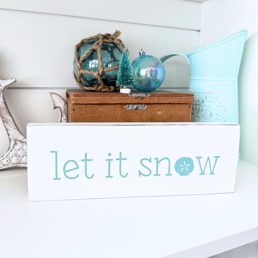 A white rectangular wooden sign with the phrase 'let it snow' in aqua blue lettering, styled on a white shelf with holiday decorations including blue ornaments, a small tree, a wooden box, and a mint pitcher.