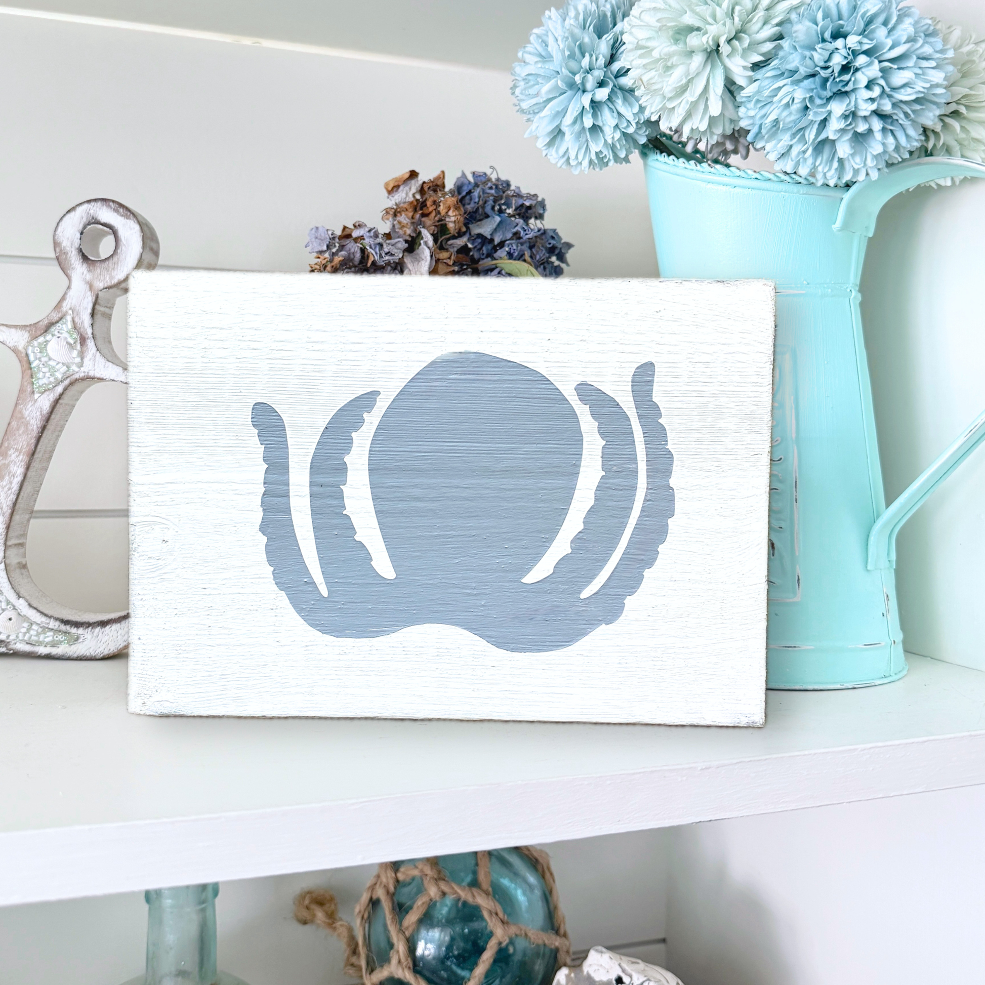 A rectangular white wooden sign with a distressed finish, featuring a painted blue octopus silhouette on the front. The sign is styled on a white shelf next to a silver anchor, dried flowers, and a light blue pitcher filled with blue pom-pom flowers.
