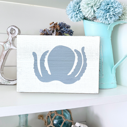 A rectangular white wooden sign with a distressed finish, featuring a painted blue octopus silhouette on the front. The sign is styled on a white shelf next to a silver anchor, dried flowers, and a light blue pitcher filled with blue pom-pom flowers.